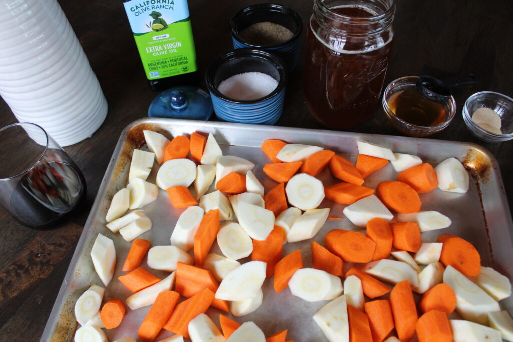 A sheet pan with seasoned parsnips and carrots waits to go in the oven. The necessary ingredients for the recipe are in the background.