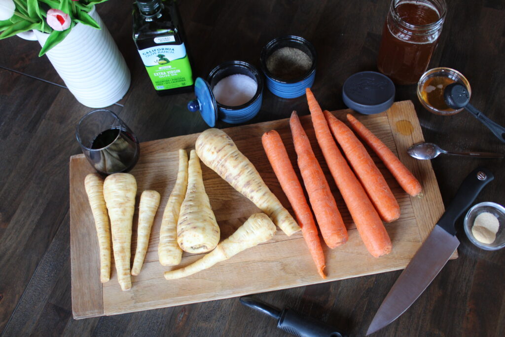Ingredients for Maple Roasted Parsnips and Carrots are laid out on a wooden cutting board to prep for the recipe. California Olive Ranch extra virgin olive oil, pure New England maple syrup, and spices are all that is needed for a simple dish. Wusthoff knife and peeler are present