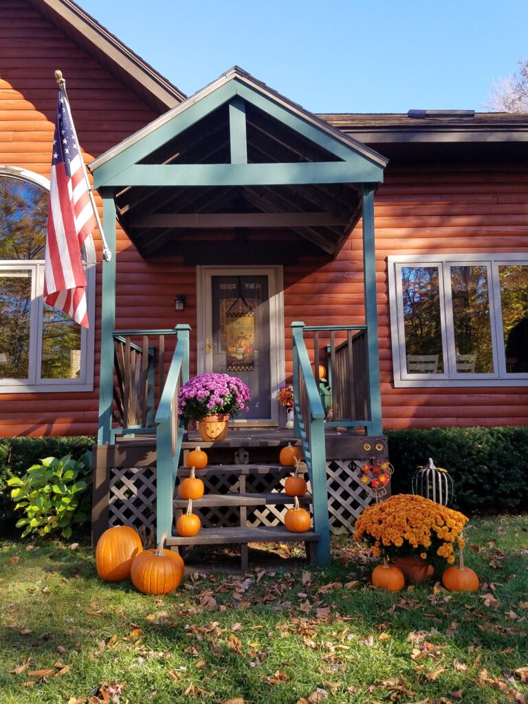 A rustic log cabin has its porch decorated with mums, pumpkins and other items for Halloween in New England
