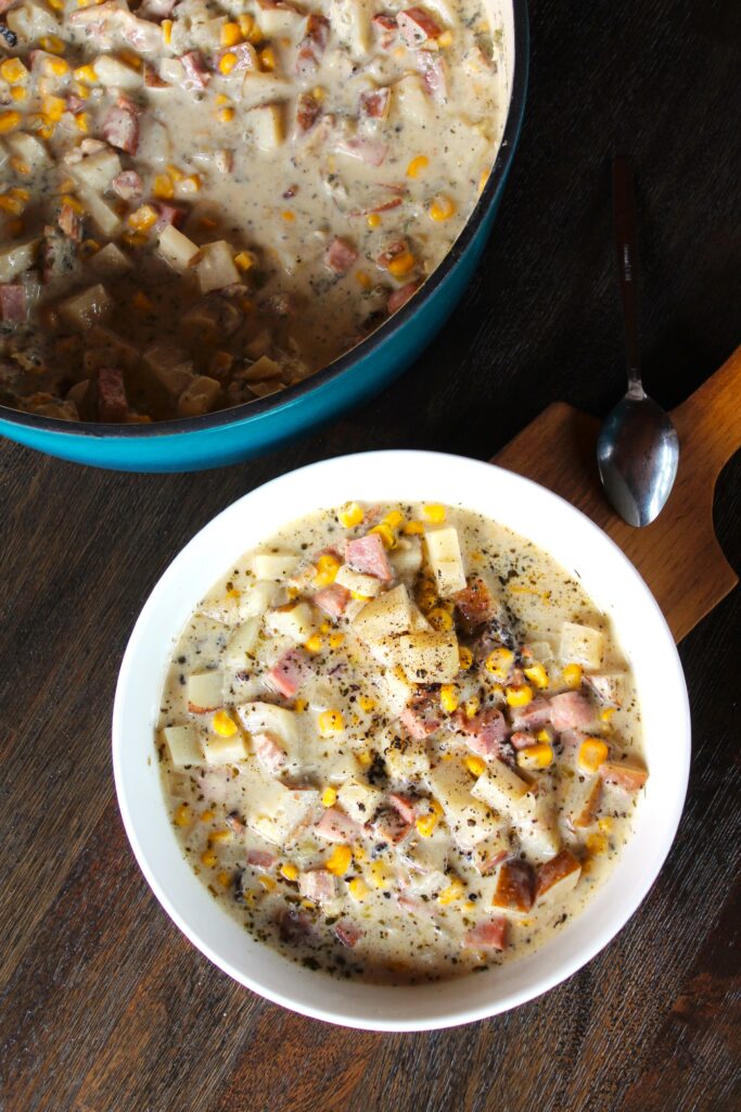 A bowl of warm ham, potato and corn chowder is dished out and ready to be served. A wide ceramic bowl shows off its beauty and color.
